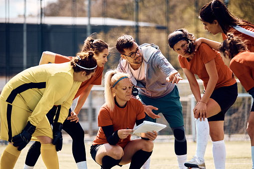 Female soccer team and their coach discussing while analyzing  game plan on playing field.
