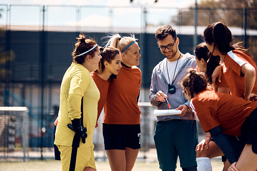 Young coach giving instructions to his female soccer team on playing field.