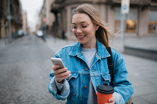 Young happy woman using a smart phone outdoors while holding a cup of coffee