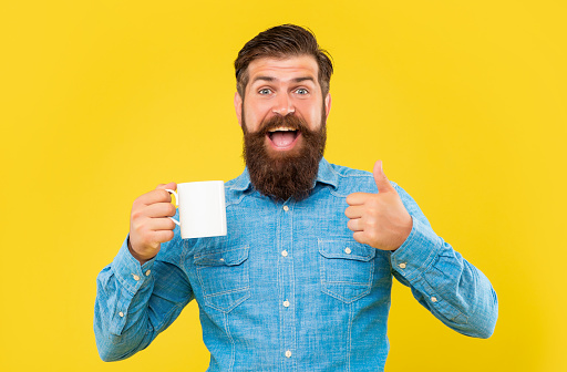 Happy man giving thumbs up holding coffee mug yellow background, copy space.
