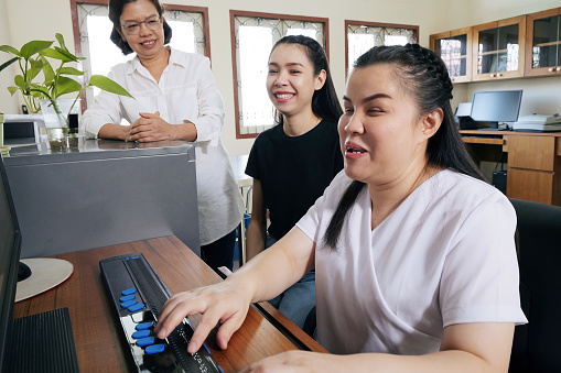 Wide angle view of happy Asian women co-workers in office workplace including person with blindness disability using computer with refreshable braille display assistive device. Disability inclusion.