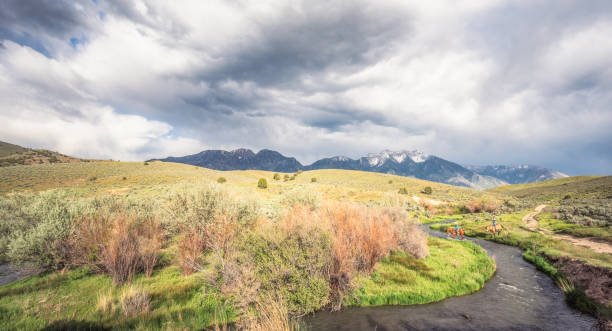A couple crossing a remote stream together on horseback, with the profile of the Rocky Mountains on the horizon.