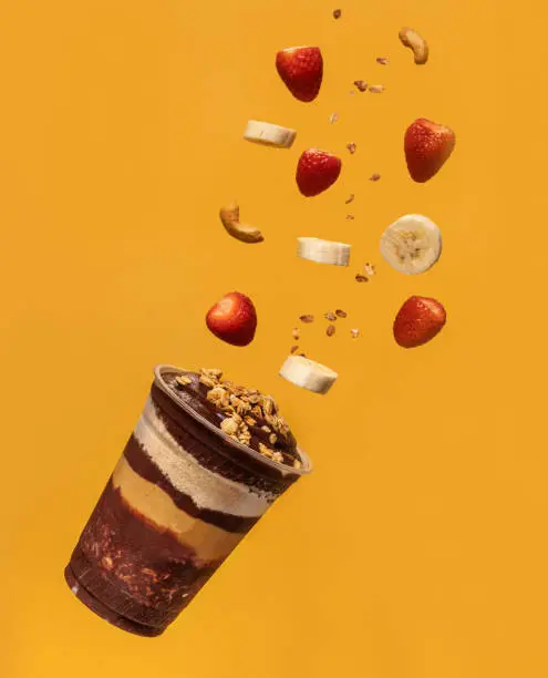 Acai cup, layers of granola, ground peanuts, and pieces of fruits and nuts flying in the air, yellow background.