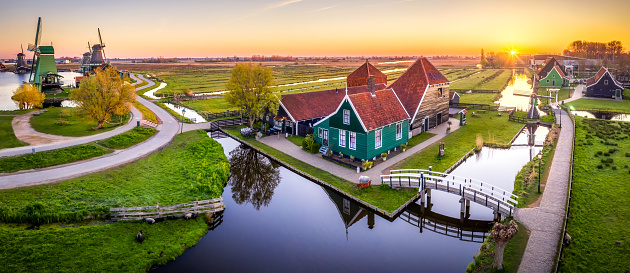 Aerial Panorama view of an antique and traditional Dutch farm houses with a wooden bridge across a canal in warm evening light. Reflection in calm water, and Dutch windmills in the background, under a clear sky with the sun rising in the morning. Location is Zaanse Schans, in Zaandam, one of the popular tourist attractions of The Netherlands.