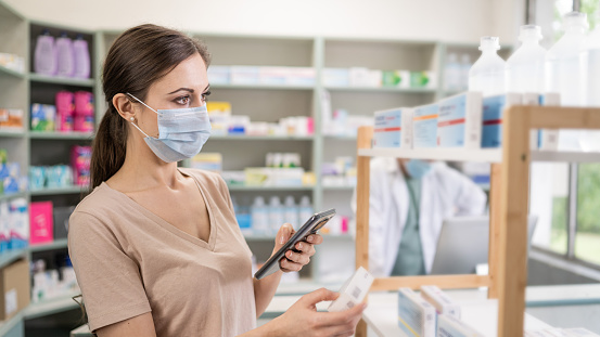 Female customer with a protective face mask in a drugstore checking online medicine with mobile phone on a patient’s  browsing aisles