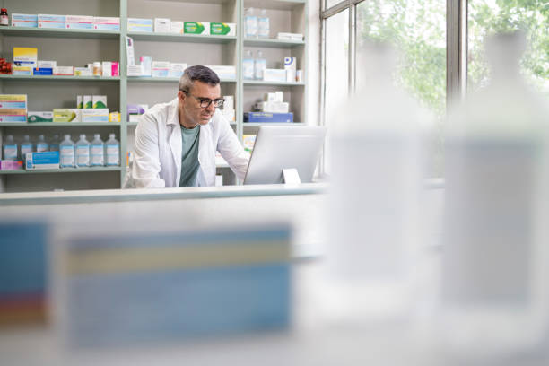 Middle age male pharmacist  checking online  medicines on a desktop computer, filing records and prescriptions, managing deliveries stock photo