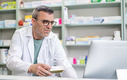 Middle age male pharmacist holding a medicine, checking online on a desktop computer, filing records and prescriptions, managing deliveries