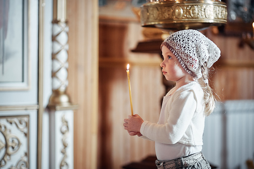 Russian little girl in a scarf on her head stands in an Orthodox Church, lights a candle and prays in front of the icon.