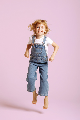 Full length view of beautiful little cheerful carefree girl in denim overalls having fun jumping barefoot on pink background.