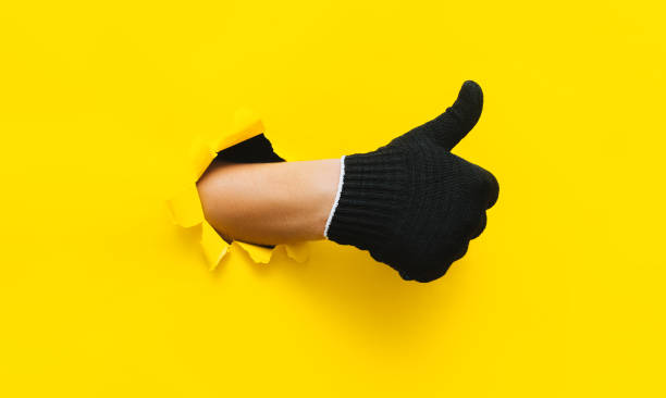 A right man's hand in a black fabric work glove shows a thumb up (like). Torn hole in yellow paper. Good job, fun mood and approval concept. Copy space. stock photo