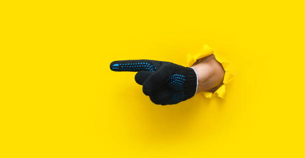 The index finger (forefinger) of a right hand in a black working knitted glove points to the left. Torn hole in yellow paper. Direction concept, copy space. stock photo