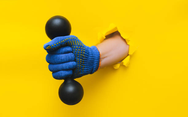 A worker's right hand in a blue knitted glove holds a black dumbbell. Torn hole in yellow paper. Healthy lifestyle concept avd copy space. stock photo