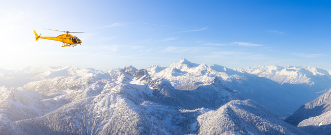 Yellow Helicopter flying over the Rocky Mountains. Winter Sunny Sky. Aerial Landscape from British Columbia, Canada near Vancouver. Adventure Composite. Canadian Panoramic Nature Background