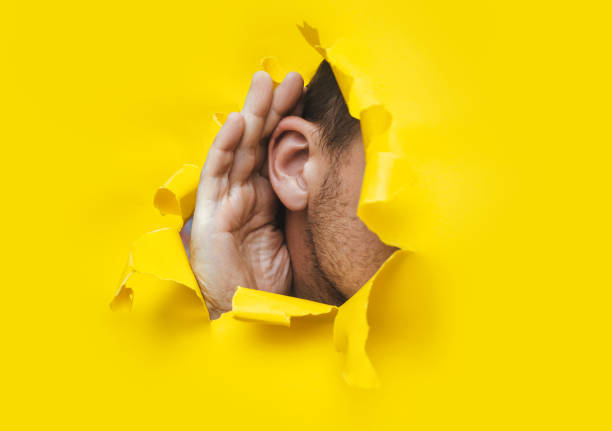 Close-up of a man's ear and hand through a torn hole in the paper. Bright yellow background, copy space. The concept of eavesdropping, espionage, gossip and tabloids. stock photo