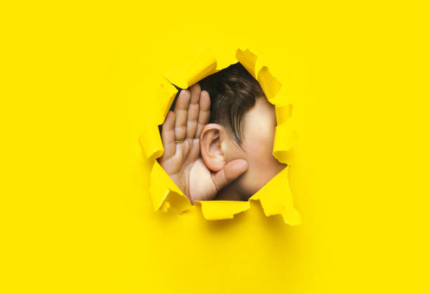 Close-up of a man's ear and hand through a torn hole in the paper. Yellow background, copy space. The concept of eavesdropping, espionage, gossip and tabloids. stock photo