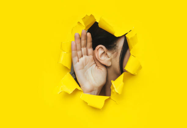 Close-up of a woman's ear and hand through a torn hole in the paper. Bright yellow background, copy space. The concept of eavesdropping, espionage, gossip and tabloids. stock photo