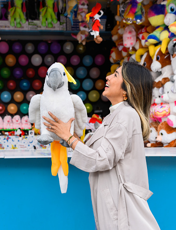 Side view of smiling female in coat with toy parrot standing near game booth with balloon darts in amusement park