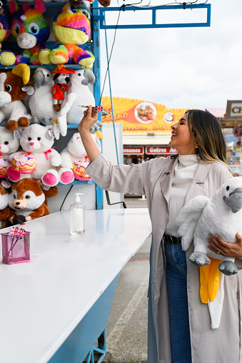 Positive female in coat with toy parrot in hand throwing dart while playing in game booth in public amusement park