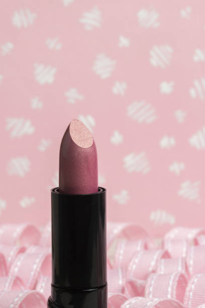 A copper pink lipstick tube standing upright, in front of rows of pastel spiral ribbon curls with a pale pink and white pattern background. Closeup with copy space.