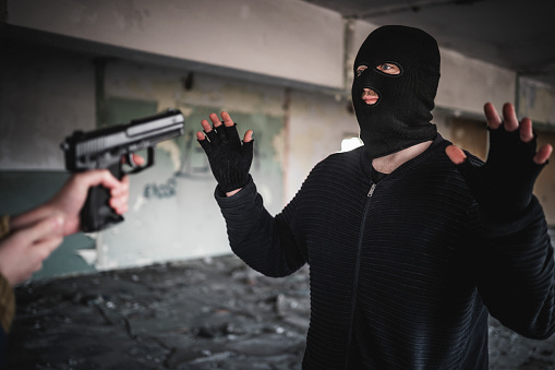 Caucasian man in black with mask with his hands up while a unrecognizable person point at him with a hand gun