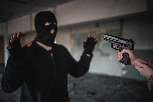 Caucasian man in black with mask with his hands up while a unrecognizable person point at him with a hand gun
