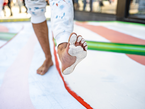 Close up of feet of Young Latin male artist painting sidewalk mural. He is dressed in casual work clothes. Exterior of public sidewalk in downtown of large North American City.