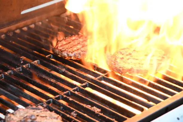 Three beef burgers being cooked on a gas fired barbecue with one burger surrounded with flames for a flamed grill burger