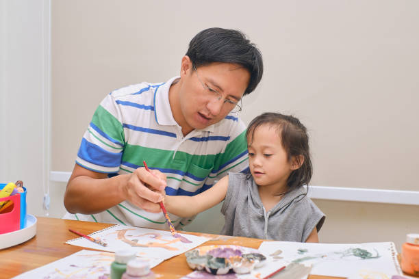 Asian father and child having fun painting with paintbrush and watercolors at home stock photo