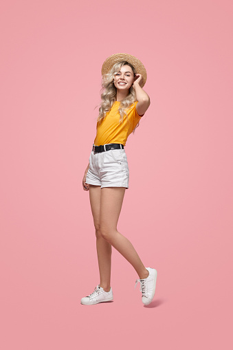 Full body of happy young female model in casual summer wear and straw hat looking at camera against pink background