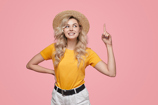Happy young blonde female in yellow t shirt and straw hat pointing up with forefinger against pink background