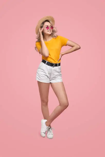 Full body young female in trendy clothes and straw hat adjusting sunglasses, and looking away with smile while holding hand on waist and standing against pink background