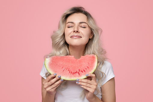 Young female with closed eyes enjoying fresh ripe sweet watermelon in summer day against pink background