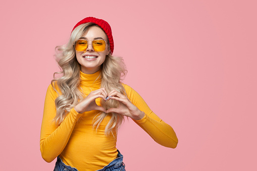 Trendy blond female in bright yellow turtleneck and glasses showing love sign and looking at camera on pink background