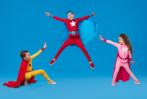 Company of preteen children in colorful costumes showing superpowers and pretending being heroes on blue background in studio