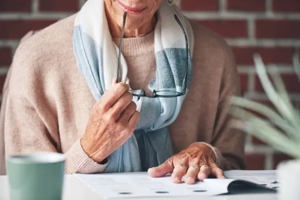 Photo of Mature woman reading financial insurance documents at home.A senior woman reading financial paperwork planning her retirement. A mature woman holding her glasses reading legal paperwork