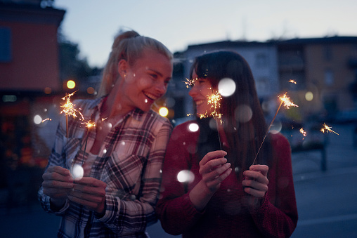 Excited friends celebrating on holiday together playing with sparklers. Happy women on holiday using fireworks to celebrate. Two young women celebrating playing with fireworks on holiday together
