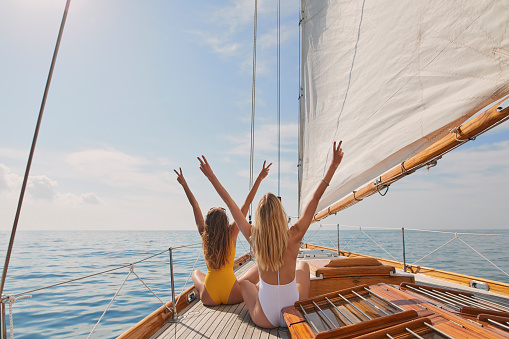 Two cheerful friends with arms raised making peace signs celebrating on boat cruise together. Two excited women in swimwear on holiday cruise celebrating cheering making peace signs in the air