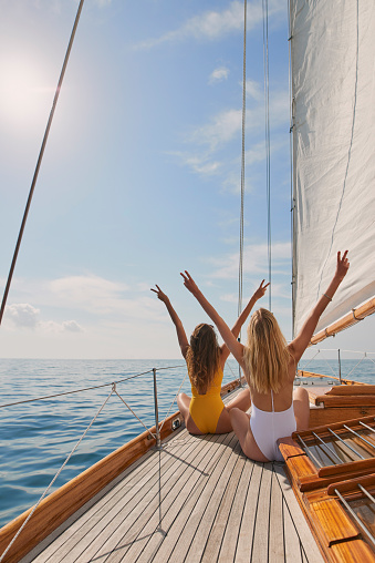 Two friends in swimsuits making peace signs celebrating on a boat cruise. Two women cheerfully celebrating making peace signs sitting on a boat. Happy friends celebrating together on a cruise