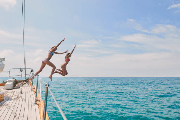 unbeschwerte aufgeregte junge frauen, die vom boot springen, um im meer zu schwimmen. zwei freunde auf einer gemeinsamen urlaubskreuzfahrt, die vom boot ins meer springen, um zu schwimmen. aufgeregte junge frauen, die gemeinsam vom boot springen - hechten stock-fotos und bilder