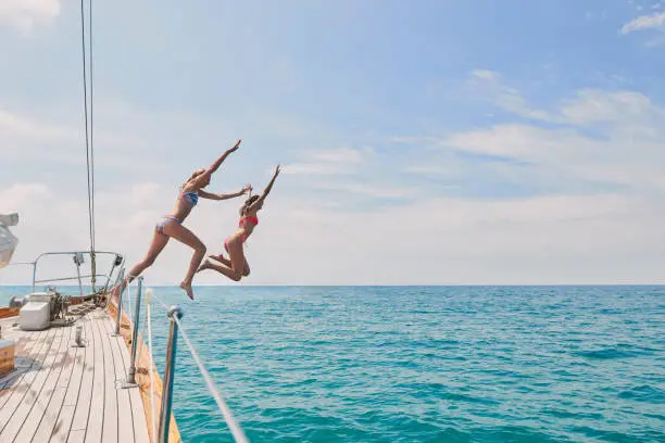 Carefree excited young women jumping from boat to swim in the ocean. Two friends on a holiday cruise together jumping from boat into the ocean to swim. Excited young women jumping off boat together