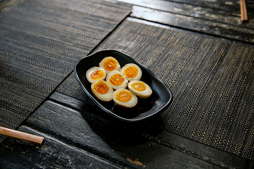 Soft-boiled Hanjuku ramen egg in the traditional Japanese ramen restaurant, with a black plate on a black mat and Japanese fonts sake barrel translation: Invincible Great Gate