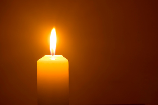 A single burning candle flame or light is glowing on a yellow candle isolated on red or dark background on table in church for Christmas, funeral or memorial service.
