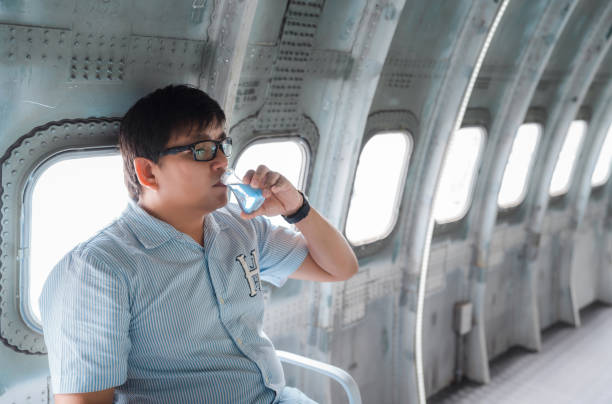 hombre asiático obeso paupérrimo bebe líquido azul de la botella de matraz erlenmeyer en un avión de acero cerca de la ventana - paunchy fotografías e imágenes de stock