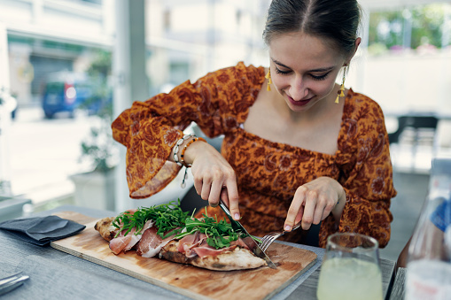 Teenage girl cutting and eating a traditional Sicilian pizza for lunch. The pizza is topped with Prosciutto di Parma and fresh arugula leaves.\nShot With Canon R5