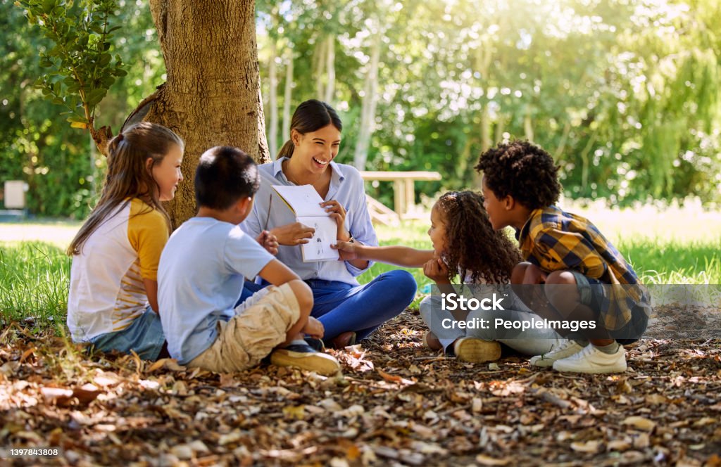 Shot of a teacher reading a book to her class in a park Time flies when you're having fun Storytelling Stock Photo