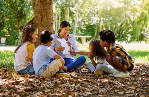 Shot of a teacher reading a book to her class in a park Time flies when you're having fun storytelling stock pictures, royalty-free photos & images