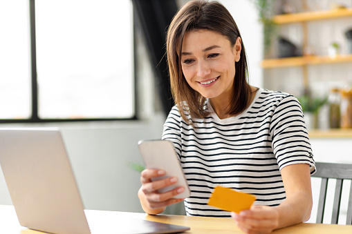 Online shopping, technology concept. Happy Caucasian woman holding mobile phone and credit card sitting at the table in the home kitchen