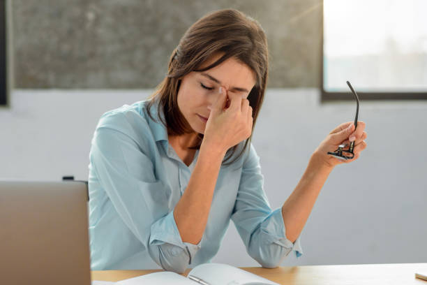 überarbeitete junge büroangestellte hält eine brille in den händen, massiert den nasenrücken, sitzt an ihrem arbeitsplatz im büro - human eye rubbing tired glasses stock-fotos und bilder