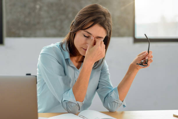 Overworked young woman office worker holds glasses in her hands, massages the bridge of her nose, sitting at her workplace in the office stock photo