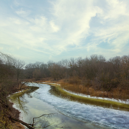 Long shot of a landscape with a small calm river that flows calmly in the winter in front of barren trees and dry grass